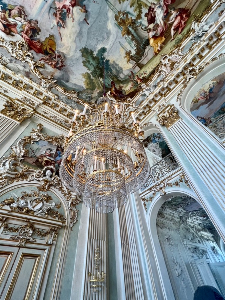 A photo of a chandelier, upper wall corner, and a painted ceiling in Schloss Nymphenburg in Munich.