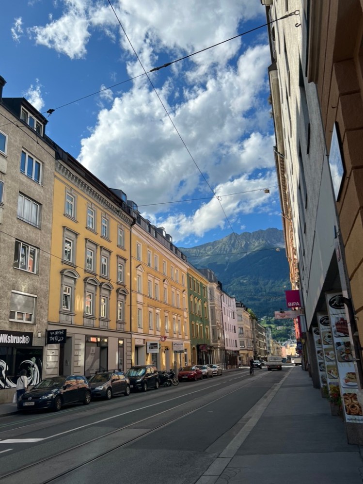 A photo of a street in Innsbruck, Austria. Building colors are pastels with mountains and a blue sky in the background.